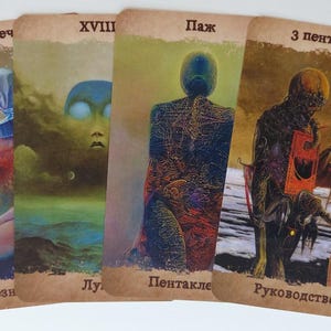 May include: Four tarot cards with surreal illustrations. The cards feature figures and landscapes in muted tones of blue, green, and brown. Each card has a title in a foreign language. The cards are arranged on a white surface.