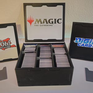 Puede incluir: Una caja de almacenamiento negra para juegos de cartas con divisores para organizar las cartas. La caja tiene tres tapas con logotipos para Magic: The Gathering, Yu-Gi-Oh! Trading Card Game y Digimon Card Game.