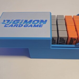 May include: A blue plastic box with the words "Digimon Card Game" on the lid. The box is open and contains a set of small, colorful, rectangular blocks.