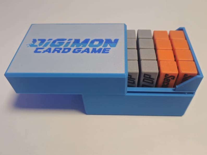 May include: A blue plastic box with the words "Digimon Card Game" on the lid. The box is open and contains a set of small, colorful, rectangular blocks.