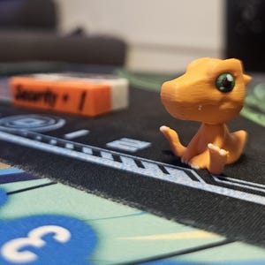 Può includere: Una piccola statuina di dinosauro arancione e bianca con occhi verdi. La statuina è seduta su una superficie nera e blu con la parola "Security" stampata su un blocco arancione e bianco vicino.