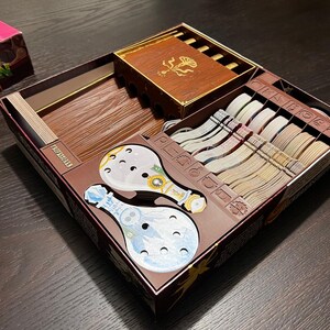 Könnte beinhalten: Eine braune Pappbox mit einem Spieleinsatz zum Organisieren von Spielteilen. Der Einsatz hat Fächer für Karten, Spielsteine und andere Spielkomponenten. Die Box ist mit "ION PROSION" beschriftet und zeigt eine rosa-grüne Illustration einer Pflanze.