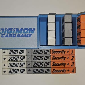 May include: A blue plastic box with a Digimon Card Game label. The box contains a set of white, gray, and orange plastic tokens with numbers and text. The tokens read: + 1000 DP + 5000 DP Security + 1, + 2000 DP + 6000 DP Security + 2, + 3000 DP + 8000 DP Security + 3, + 4000 DP + 10000 DP Security + 4.