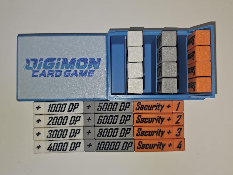 May include: A blue plastic box with a Digimon Card Game label. The box contains a set of white, gray, and orange plastic tokens with numbers and text. The tokens read: + 1000 DP + 5000 DP Security + 1, + 2000 DP + 6000 DP Security + 2, + 3000 DP + 8000 DP Security + 3, + 4000 DP + 10000 DP Security + 4.