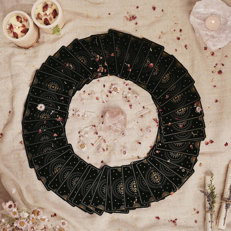 May include: A circular arrangement of tarot cards with a celestial design on a cream-colored surface. A pink crystal and clear quartz crystals are in the center. Dried flowers and two candles with rose petals surround the cards, creating a mystical aesthetic.