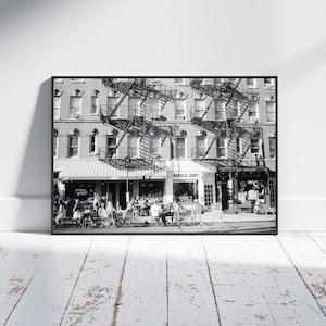 Puede incluir: Fotografía en blanco y negro de una escena callejera de la ciudad de Nueva York. La imagen presenta un edificio de varios pisos con escaleras de incendios, una barbería y personas sentadas en mesas al aire libre. La fotografía está enmarcada y expuesta en una pared blanca.