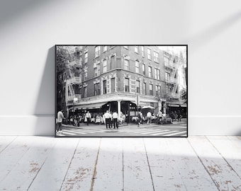 May include: Black and white framed print of a New York City street scene. The image features a multi-story brick building with a restaurant on the corner. People are dining at tables outside. Fire escapes are visible on the building.