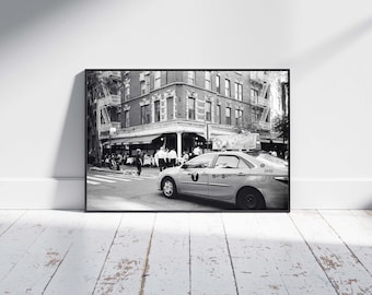 May include: Black and white framed photograph of a New York City street scene. A yellow taxi cab is parked on the street in front of a restaurant. People are walking on the sidewalk and crossing the street.