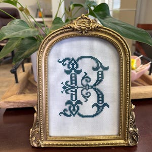 May include: A gold-colored ornate frame with a white background and a green embroidered letter "B" in a decorative font.
