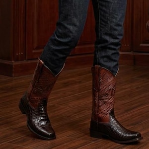 Op de afbeelding: Een bruine cowboyhoed, een wit overhemd met lange mouwen en knopen, een donkerbruin leren vest met vijf knopen, een donkerblauwe spijkerbroek en bruine leren cowboylaarzen met een textuur op het onderste deel en decoratieve stiksels.