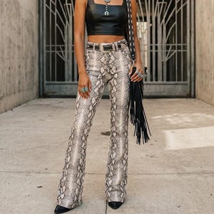 May include: A black sleeveless faux leather crop top paired with high-waisted flared trousers featuring a grey and black snakeskin print. The outfit includes a black studded belt with a silver buckle and a black shoulder bag with long fringe details.
