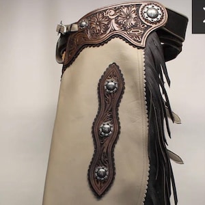 May include: Taupe leather chaps with dark brown and black accents. The chaps feature decorative silver studs, intricate tooling, and a fringe along the edge. The text "Taupe Chinks" is visible at the bottom.