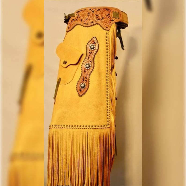 K&ouml;nnte beinhalten: Senfgelbe Lederchaps mit aufwendigen Werkzeugen und Fransen. Die Chaps verf&uuml;gen &uuml;ber eine dekorative Lederverkleidung mit silberfarbenen Nieten und einer kleinen Tasche. Der G&uuml;rtel ist ebenfalls mit floralen Werkzeugen verziert.