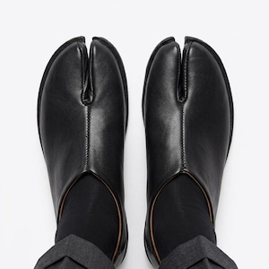 Pode incluir: Um par de sapatos pretos de couro com biqueira dividida, também conhecidos como sapatos Tabi, com um design distinto. Os sapatos têm uma estética elegante e minimalista, com uma costura visível no centro do dedo do pé. Os sapatos são usados com meias pretas e calças cinzentas.