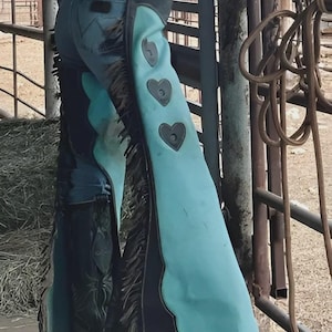 May include: Light blue cowboy chaps with black trim and heart-shaped embellishments. The chaps, with a fringed edge, are worn over denim jeans. The image evokes a western aesthetic.
