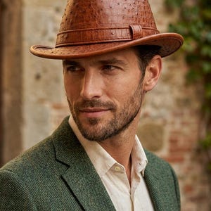 Puede incluir: Un sombrero fedora marrón con un patrón texturizado de piel de avestruz y una banda de cuero a juego alrededor de la copa. El sombrero presenta una copa pellizcada y un ala curva, mostrando una textura superficial detallada con pequeños relieves.