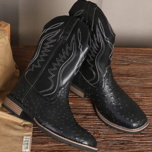 Puede incluir: Un par de botas de vaquero negras con punta cuadrada y costuras blancas decorativas. Las botas tienen un aspecto de cuero texturizado y una suela marrón claro. Las botas se exhiben sobre una superficie de madera.