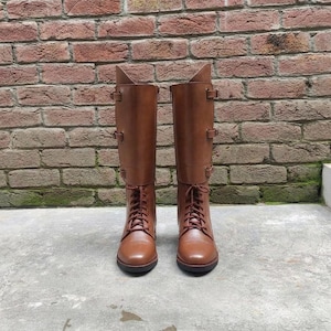 Könnte beinhalten: Ein Paar kniehohe braune Stiefel mit Schnürung vorne und passenden braunen Schnürsenkeln. Jeder Stiefel verfügt über einen hohen Schaft mit drei horizontalen Riemen und Schnallen an der Außenseite, eine abgerundete Zehenkappe und eine flache Sohle.