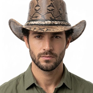 Puede incluir: Un sombrero de vaquero con un diseño de piel de serpiente en tonos marrones y negros. El sombrero tiene un ala ancha y una copa alta. Una banda decorativa rodea la base de la copa. La persona lleva una camisa verde.