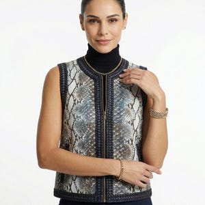 May include: A sleeveless vest with a snakeskin pattern in shades of gray, blue, and white. The vest has a dark blue trim and a zipper closure. The person is wearing a black turtleneck, gold necklace, and gold bracelets.