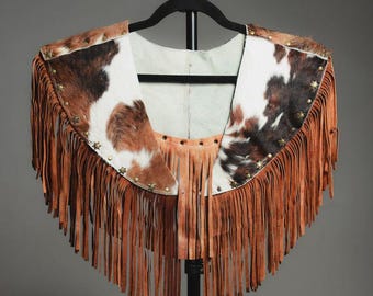 May include: A western-style leather cape with brown fringe and cowhide accents. The cape features a white and brown cowhide pattern on the shoulders and is adorned with star-shaped studs. The fringe hangs from the bottom edge.