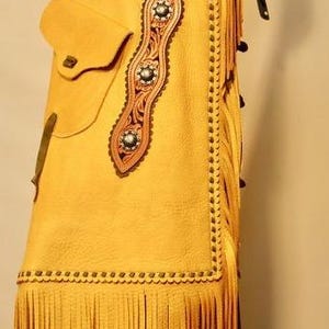 K&ouml;nnte beinhalten: Senfgelbe Lederchaps mit aufwendigen floralen Verzierungen aus Leder. Merkmale sind eine Tasche, dekorative N&auml;hte und lange Fransen am unteren Rand. Die Chaps haben einen dekorativen G&uuml;rtel mit floralem Design.