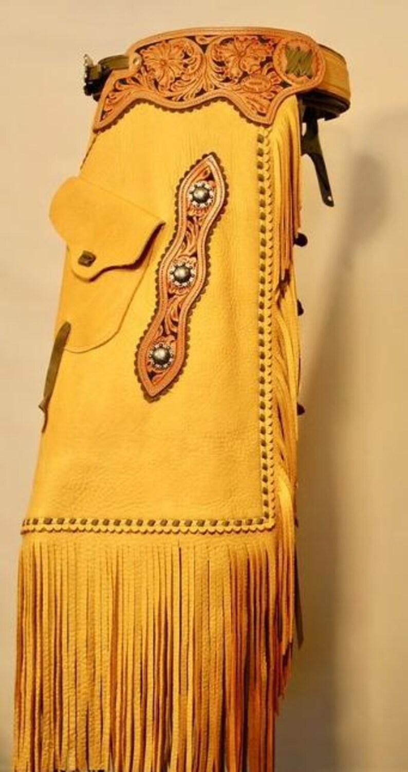 K&ouml;nnte beinhalten: Senfgelbe Lederchaps mit aufwendigen floralen Verzierungen aus Leder. Merkmale sind eine Tasche, dekorative N&auml;hte und lange Fransen am unteren Rand. Die Chaps haben einen dekorativen G&uuml;rtel mit floralem Design.