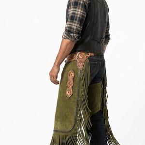 Pode incluir: Chaps de camurça verde-oliva com franjas longas e detalhes decorativos em couro. Os chaps são usados com um colete de couro preto, camisa xadrez e jeans. Roupas ocidentais.