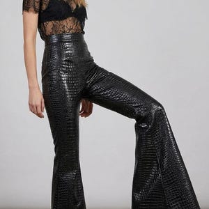 Könnte beinhalten: Ein schwarzes kurzärmeliges Spitzenoberteil mit Rundhalsausschnitt und einem schwarzen Bralette darunter, kombiniert mit einer schwarzen Hose mit hoher Taille, Krokodilleder-Strukturmuster und einem bis zum Boden reichenden Schlag.