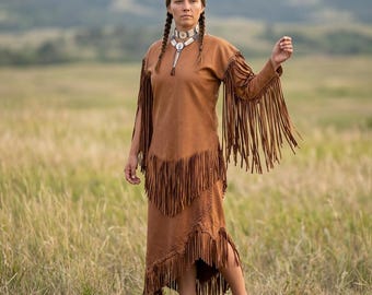 May include: A tan suede dress featuring long sleeves with extensive fringe detailing, a layered fringe bodice, and a tiered fringe skirt. The garment includes a round neckline and is paired with a silver-toned metal choker necklace.