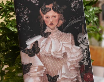 Peut inclure: Une carte de tarot représentant une femme vêtue d'une robe blanche à volants et d'une couronne de papillons. La carte est intitulée "La Mort" et a un fond sombre avec des fleurs et des crânes.