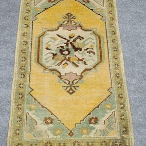 Puede incluir: Una alfombra rectangular que presenta un medallón central en forma de diamante amarillo con un interior color crema que contiene motivos florales marrones y verdes. La alfombra tiene un borde verde con patrones marrones y flecos en ambos extremos.