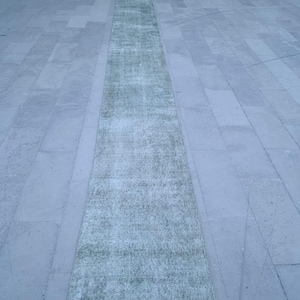 May include: A rectangular, distressed-look rug in shades of green and gray. The rug is placed on a light gray stone surface, with a visible border. The rug's design appears faded, giving it a vintage aesthetic. The rug is long and narrow, suitable for a hallway or entryway.