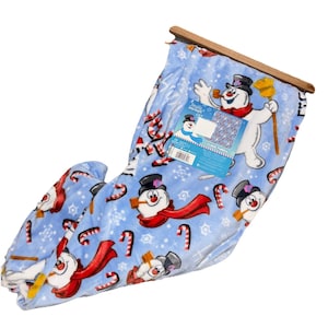 May include: A light blue blanket featuring a repeating pattern of snowmen with black top hats, red scarves, and candy canes. The blanket is held by a wooden bar at the top and has a tag attached with the words "Frosty Snowman".