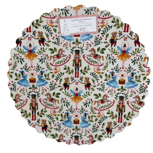 May include: A round, white placemat with a scalloped edge, featuring a festive pattern of nutcrackers, ballerinas, mice, and rocking horses. The design includes green foliage and red berries. The placemat has a tag that reads "cupcakes and cashmere".