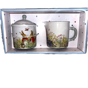 May include: A ceramic sugar bowl with a lid and a creamer with a handle, both decorated with a floral and bunny rabbit design. The set is presented in a decorative box with a blue border and polka dots.