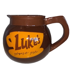 May include: A brown ceramic mug with a large handle. The mug features the text "Luke's" in brown letters on a yellow background, with "gilmore girls" written below. The mug has a rounded shape.