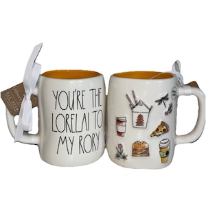 May include: Two white ceramic mugs with yellow interiors. One mug has the text "YOU'RE THE LORELAI TO MY RORY" in black. The other mug is decorated with food-themed stickers, including a pizza slice and a burger. Each mug has a white ribbon attached.