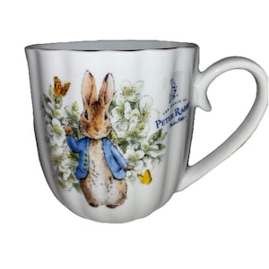 May include: White ceramic mug featuring Peter Rabbit in a blue jacket, surrounded by flowers and butterflies. The mug has a gold rim and a curved handle. Text on the mug reads "The World of Peter Rabbit".