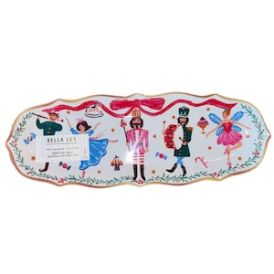 May include: A white serving tray with a gold trim and a festive Nutcracker theme. The tray features illustrations of a ballerina, a drummer, a fairy, and a nutcracker soldier. The tray has the text "BELLA LUX" and "SERVING TRAY".