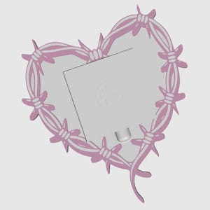 May include: A pink heart-shaped frame with a barbed wire design. The frame surrounds a light gray rectangular space, possibly for a photo or artwork. The design is a mix of soft and sharp elements.