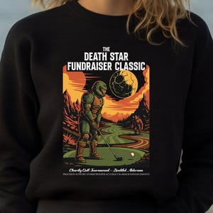 May include: Black sweatshirt featuring a colorful graphic of a figure in armor playing golf on a green course under a Death Star. The text reads "THE DEATH STAR FUNDRAISER CLASSIC". Additional text at the bottom mentions a charity golf tournament.