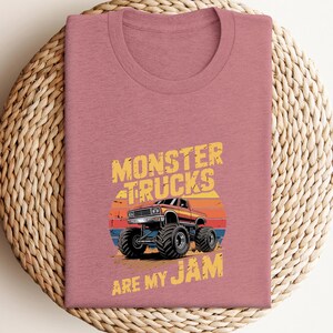 May include: A dusty rose t-shirt featuring a graphic of a monster truck with the text "MONSTER TRUCKS ARE MY JAM". The design includes a sunset backdrop with orange, red, and blue stripes. The shirt is folded on a woven, circular placemat.