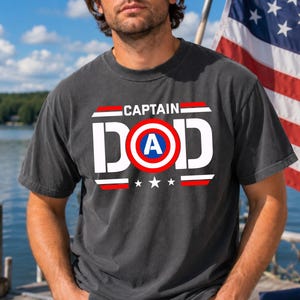 K&ouml;nnte beinhalten: Dunkelgraues T-Shirt mit dem Schriftzug "CAPTAIN DAD" in Wei&szlig;, Rot und Blau. Das Design beinhaltet ein Schild mit dem Buchstaben "A" darin und drei Sternen darunter. Das T-Shirt wird von einer Person getragen.
