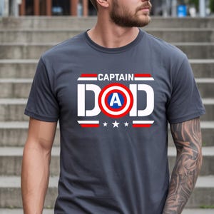 K&ouml;nnte beinhalten: Graues T-Shirt mit dem Text "CAPTAIN DAD" in Wei&szlig;, Rot und Blau. Das Design beinhaltet ein Schild mit dem Buchstaben "A" und drei Sternen unter dem Wort "DAD".
