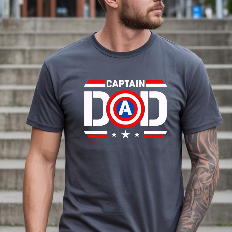 K&ouml;nnte beinhalten: Graues T-Shirt mit dem Text "CAPTAIN DAD" in Wei&szlig;, Rot und Blau. Das Design beinhaltet ein Schild mit dem Buchstaben "A" und drei Sternen unter dem Wort "DAD".