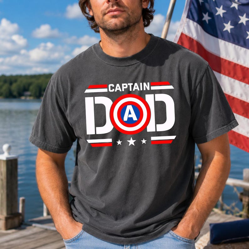 K&ouml;nnte beinhalten: Dunkelgraues T-Shirt mit dem Schriftzug "CAPTAIN DAD" in Wei&szlig;, Rot und Blau. Das Design beinhaltet ein Schild mit dem Buchstaben "A" darin und drei Sternen darunter. Das T-Shirt wird von einer Person getragen.
