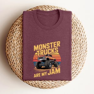 May include: Maroon t-shirt featuring a graphic of a monster truck with the text "MONSTER TRUCKS ARE MY JAM". The design includes a sunset backdrop. The shirt is folded on a woven, circular surface.