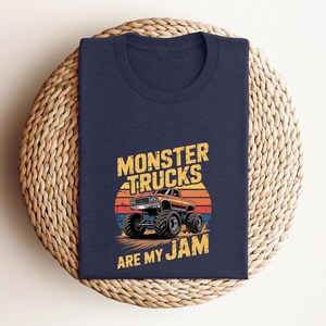May include: Navy blue t-shirt featuring a graphic of a monster truck with the text "MONSTER TRUCKS ARE MY JAM". The design includes a sunset backdrop in shades of orange, red, and blue. The shirt is folded on a woven surface.