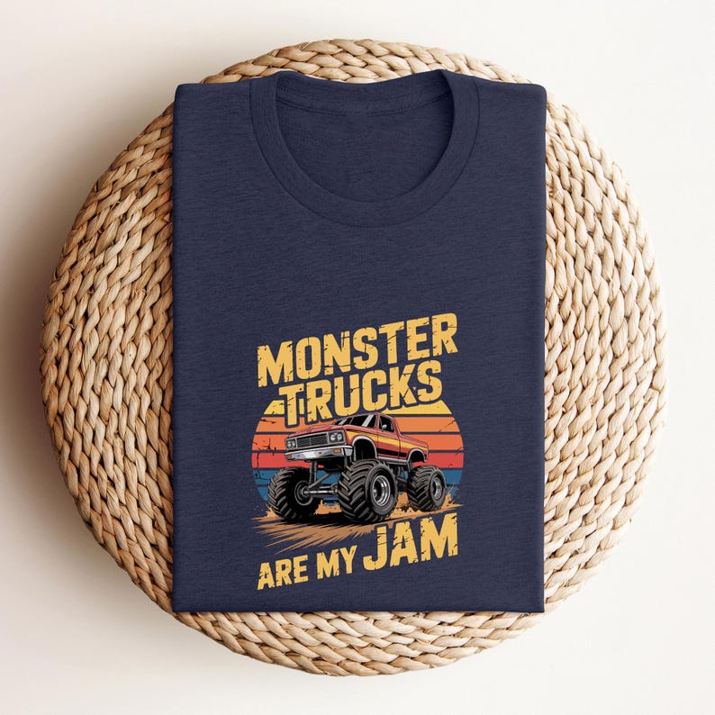 May include: Navy blue t-shirt featuring a graphic of a monster truck with the text "MONSTER TRUCKS ARE MY JAM". The design includes a sunset backdrop in shades of orange, red, and blue. The shirt is folded on a woven surface.